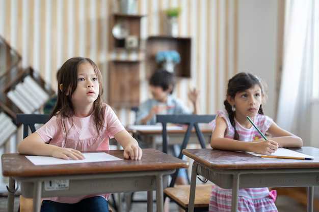 Two young girls at desks looking attentive while writing, with classmates studying in the background.

